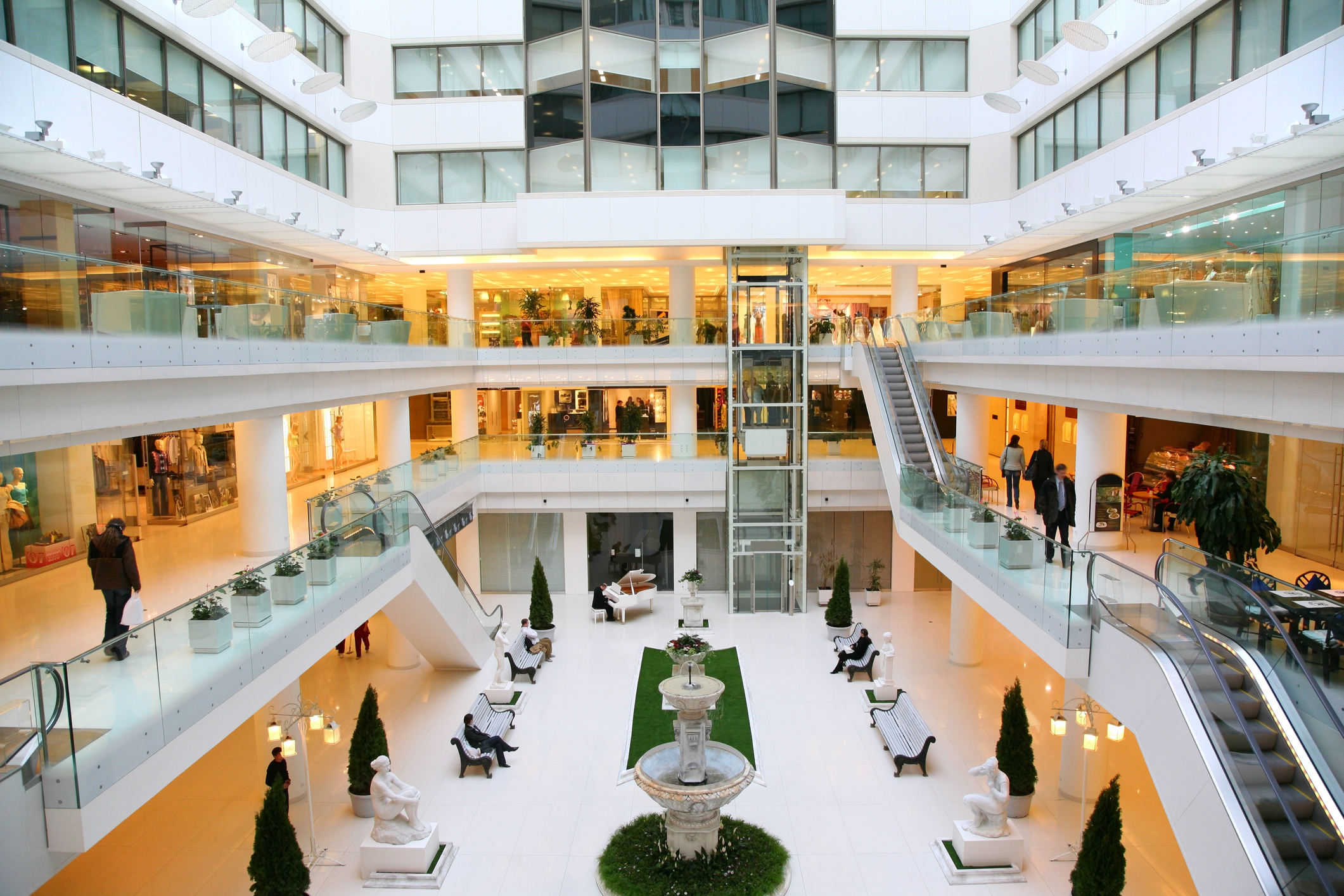 A spacious multi-level shopping center with glass railings, escalators, a central courtyard with a fountain, benches, and decorative statues.