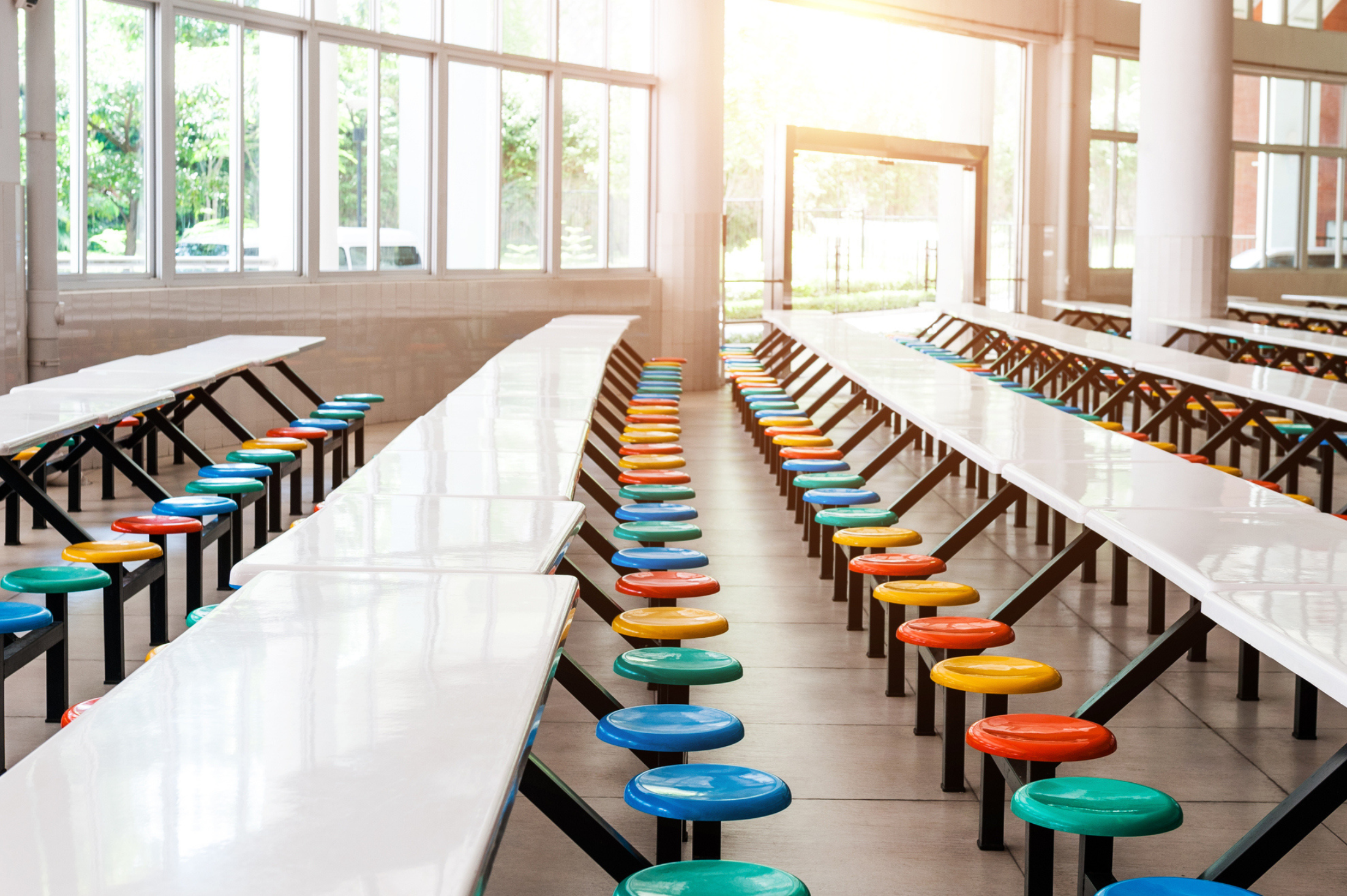 A brightly lit cafeteria with long white tables and colorful round stools arranged in neat rows.