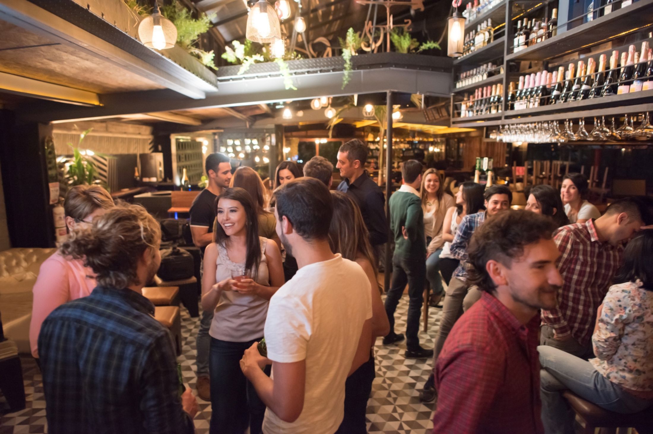 A lively bar with a warm atmosphere, dim lighting, hanging plants, and groups of people engaged in conversation.