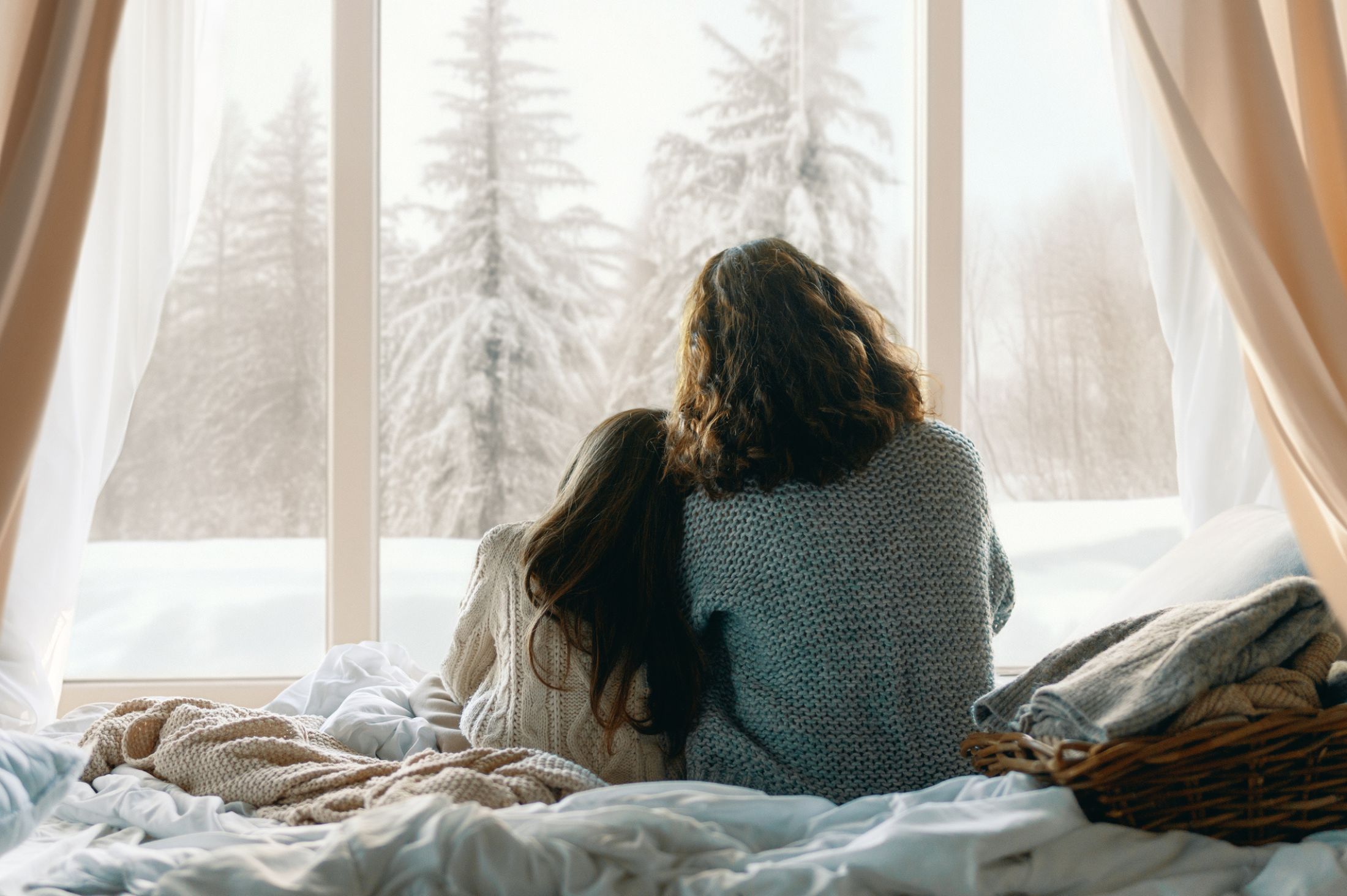 A woman and child sit on a cozy bed near a large window, wrapped in knitted sweaters, looking out at a snowy winter landscape.